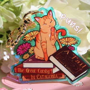 Op de afbeelding: Een heldere acryl sleutelhanger met een oranje tabby kat die op een stapel boeken zit. De boeken heten "The Great Catsby in Catagonia" en "A Tail of Two Kitties". Het woord "puns!" staat boven de kat.