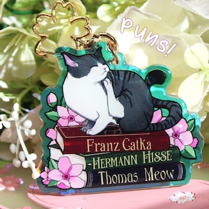 Könnte beinhalten: Eine schwarz-weiße Katze mit grünen Augen sitzt auf einem Stapel Bücher. Die Bücher tragen die Titel "Franz Catka", "Hermann Hisse" und "Thomas Meow". Die Katze ist von rosa Blumen und grünen Blättern umgeben. Der Hintergrund ist ein verschwommenes Bild von weißen Blumen.