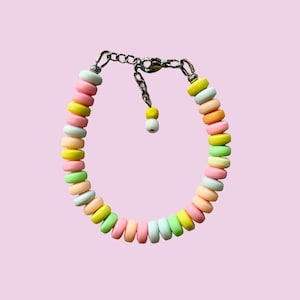 Könnte beinhalten: Ein farbenfrohes Armband mit pastellfarbenen, kreisförmigen Perlen in Rosa, Gelb, Grün und Orange. Das Armband ist an einer silberfarbenen Kette mit einem Karabinerverschluss befestigt. Der Hintergrund ist hellrosa.
