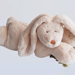 Pode incluir: Um coelho de pelúcia de cor creme com orelhas compridas e caídas, deitado em um travesseiro verde e marrom. O coelho tem um nariz costurado e os olhos fechados.