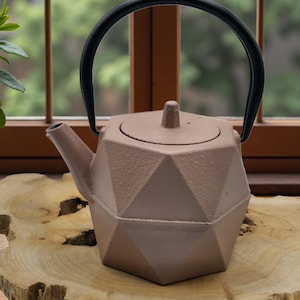 Op de afbeelding: Een geometrische, lichtbruine theepot met een zwarte handgreep. De theepot heeft een textuuroppervlak en een klein deksel. De tuit loopt naar links. De theepot staat op een houten oppervlak.