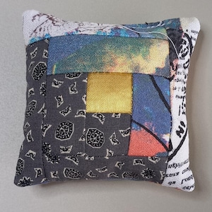 May include: A square pin cushion with a patchwork design. The cushion features a mix of fabrics in various colors and patterns, including gray, blue, yellow, and red. The edges are finished with a white fabric printed with text.