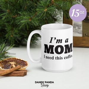 Op de afbeelding: Witte keramische mok van 443 ml met de zwarte tekst "I'm a MOM I need this coffee". De mok heeft een groot handvat. De achtergrond omvat groen en een houten kist.