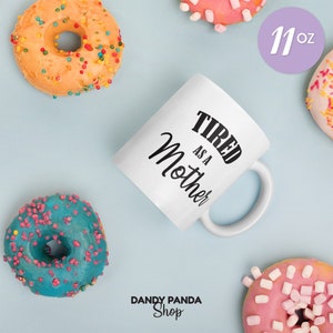 Op de afbeelding: Witte keramische mok met de tekst "TIRED AS A Mother" in zwart schrift. De mok is omringd door kleurrijke donuts met hagelslag en glazuur. De mok is 312 gram.