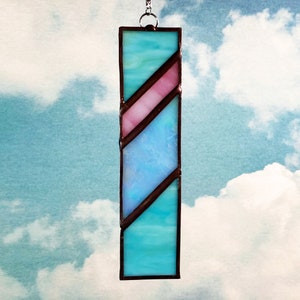 May include: A stained glass suncatcher with a blue and pink design. The suncatcher is rectangular and has a brown metal frame.