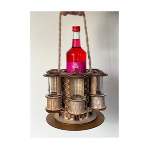 Op de afbeelding: Een houten drankdrager met een fles rode vloeistof en verschillende shotglaasjes. De drager heeft een ronde basis en een handvat van touw. De fles heeft een rode dop en een etiket. De drager is gemaakt van lichtbruin hout.
