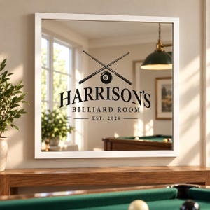 Op de afbeelding: Een vierkante wandspiegel met een witte lijst en een zwarte afbeelding van twee gekruiste biljartkeus en een achtbal. De tekst luidt HARRISON'S BILLIARD ROOM EST. 2026. De spiegel reflecteert een kamer met een biljarttafel en een groene hanglamp.