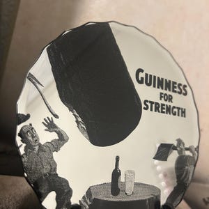 May include: A white ceramic plate with a black and white illustration featuring the text "Guinness For Strength." The plate depicts a scene with figures, a bottle, and a glass. The plate has a wavy edge.