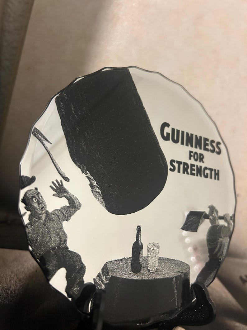 May include: A white ceramic plate with a black and white illustration featuring the text "Guinness For Strength." The plate depicts a scene with figures, a bottle, and a glass. The plate has a wavy edge.