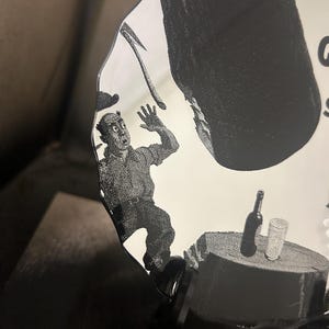 May include: A black and white illustration on a decorative mirror. The image depicts a man in a state of panic, with a large object falling towards him. A table with a bottle and glass is also visible.