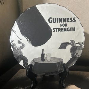 May include: A black and white Guinness advertisement on a decorative plate. The plate features a cartoon illustration of two men, a table with a bottle and glass, and the text "Guinness For Strength."