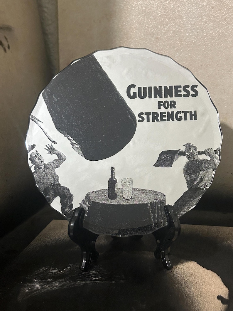 May include: A black and white Guinness advertisement on a decorative plate. The plate features a cartoon illustration of two men, a table with a bottle and glass, and the text "Guinness For Strength."