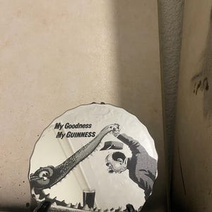 Op de afbeelding: Decoratieve zwart-wit plaat met de tekst "My Goodness My GUINNESS". De plaat toont een cartoon illustratie van een wezen en een man die naar een pint Guinness grijpt.