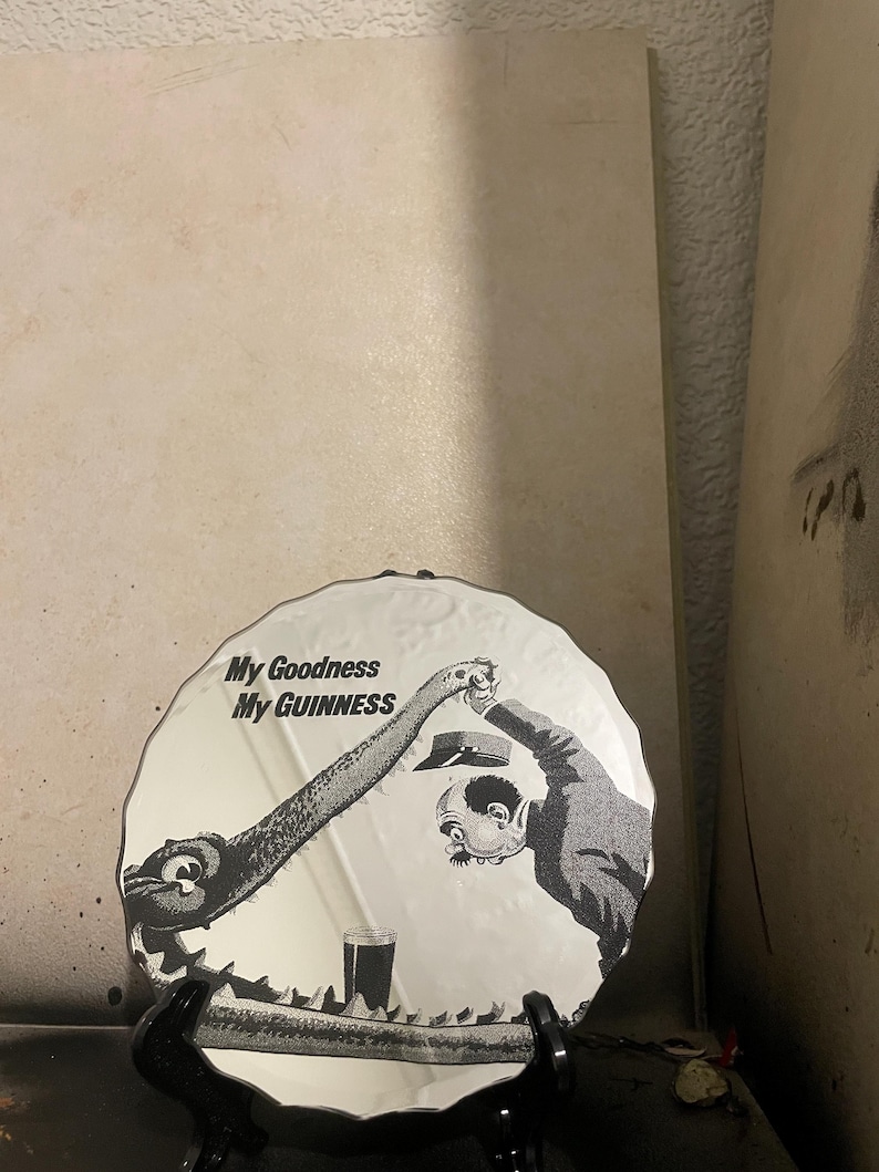 Op de afbeelding: Decoratieve zwart-wit plaat met de tekst "My Goodness My GUINNESS". De plaat toont een cartoon illustratie van een wezen en een man die naar een pint Guinness grijpt.