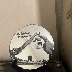 Op de afbeelding: Ronde decoratieve spiegel met een zwart-wit illustratie en de tekst "My Goodness My Guinness". De afbeelding toont een cartoonsc&egrave;ne met een Guinness-glas.