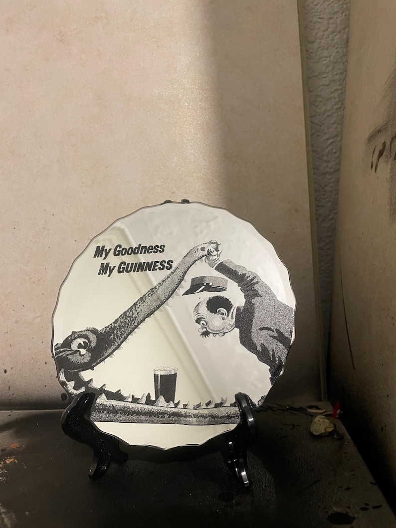 Op de afbeelding: Ronde decoratieve spiegel met een zwart-wit illustratie en de tekst "My Goodness My Guinness". De afbeelding toont een cartoonsc&egrave;ne met een Guinness-glas.