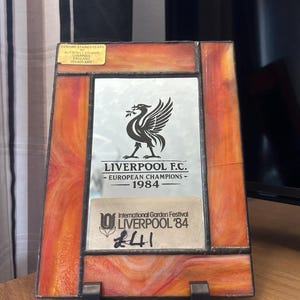 Può includere: Una targa rettangolare in vetro colorato con bordi in vetro marmorizzato arancione e rosso che circondano uno specchio centrale. Lo specchio presenta un logo nero del Liverpool F.C., il testo "LIVERPOOL F.C. EUROPEAN CHAMPIONS 1984" e un adesivo per l'International Garden Festival Liverpool '84.