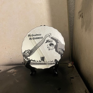Op de afbeelding: Een witte decoratieve plaat met een zwart-wit illustratie van een stripfiguur die een drankje inschenkt. De plaat heeft de tekst "My Goodness My Guinness". Het wordt op een zwarte standaard getoond.