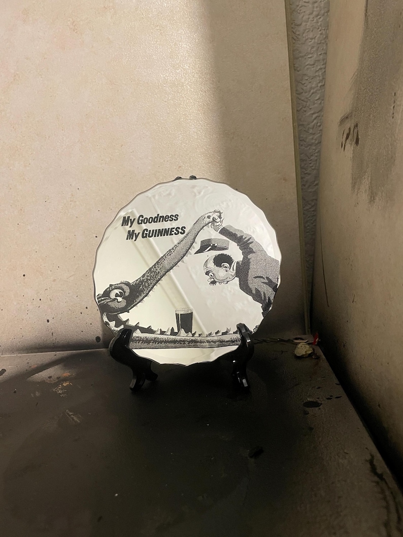 Op de afbeelding: Een witte decoratieve plaat met een zwart-wit illustratie van een stripfiguur die een drankje inschenkt. De plaat heeft de tekst "My Goodness My Guinness". Het wordt op een zwarte standaard getoond.