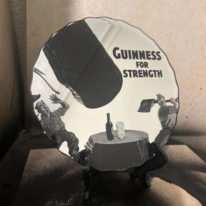 May include: A black and white Guinness advertisement on a round, decorative mirror. The image depicts a large black object falling on a table with a bottle and glass, with the text "Guinness for Strength". The mirror is displayed on a black stand.