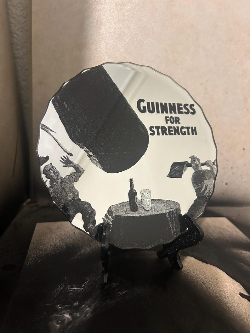 May include: A black and white Guinness advertisement on a round, decorative mirror. The image depicts a large black object falling on a table with a bottle and glass, with the text "Guinness for Strength". The mirror is displayed on a black stand.