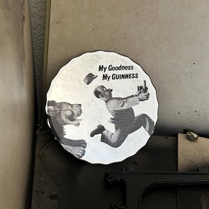 K&ouml;nnte beinhalten: Rundes Schwarz-Wei&szlig;-Grafik mit dem Text "My Goodness My Guinness". Das Bild zeigt einen Cartoon-Mann, der vor einem L&ouml;wen flieht und eine Flasche Guinness h&auml;lt. Der L&ouml;we verfolgt den Mann.