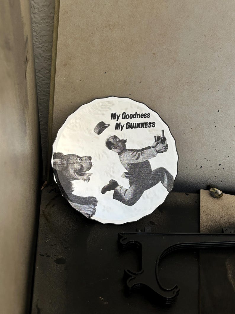 K&ouml;nnte beinhalten: Rundes Schwarz-Wei&szlig;-Grafik mit dem Text "My Goodness My Guinness". Das Bild zeigt einen Cartoon-Mann, der vor einem L&ouml;wen flieht und eine Flasche Guinness h&auml;lt. Der L&ouml;we verfolgt den Mann.