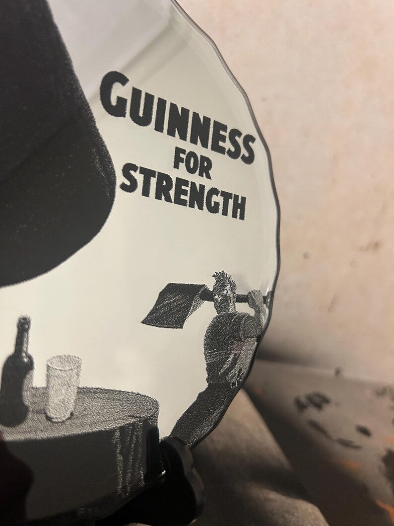 May include: A clear, decorative glass plate with the words "Guinness For Strength" in bold black letters. The plate features a black and white illustration of a man swinging a shovel, with a bottle and glass on a table.