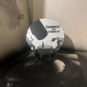 May include: A black and white Guinness advertisement on a round plate. The plate features the text "Guinness for Strength" and an illustration of a table with a bottle and glass. The plate is displayed on a black stand.