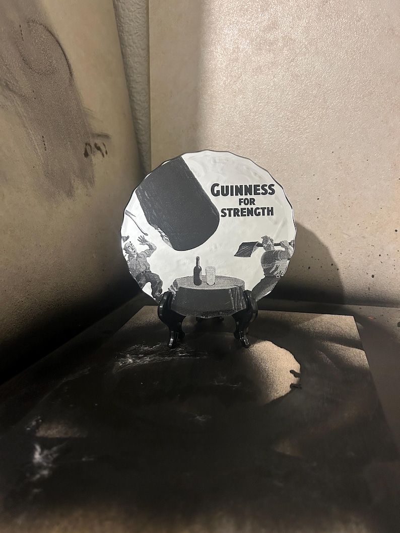 May include: A black and white Guinness advertisement on a round plate. The plate features the text "Guinness for Strength" and an illustration of a table with a bottle and glass. The plate is displayed on a black stand.