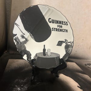 May include: A decorative, round mirror with a black and white Guinness advertisement. The image features two men, a table with a bottle and glass, and the text "Guinness FOR STRENGTH". The mirror is displayed on a black stand.