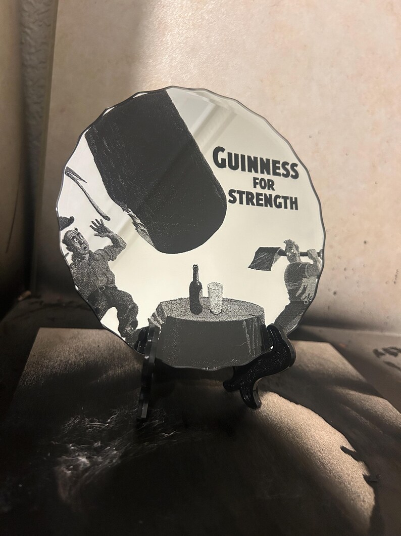 May include: A decorative, round mirror with a black and white Guinness advertisement. The image features two men, a table with a bottle and glass, and the text "Guinness FOR STRENGTH". The mirror is displayed on a black stand.