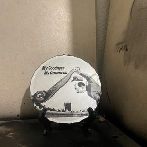 Op de afbeelding: Een zwart-wit decoratief bord met de tekst "My Goodness My Guinness". Het bord toont een cartoonillustratie van twee figuren die toosten met een pint Guinness. Het bord wordt op een zwarte standaard tentoongesteld.