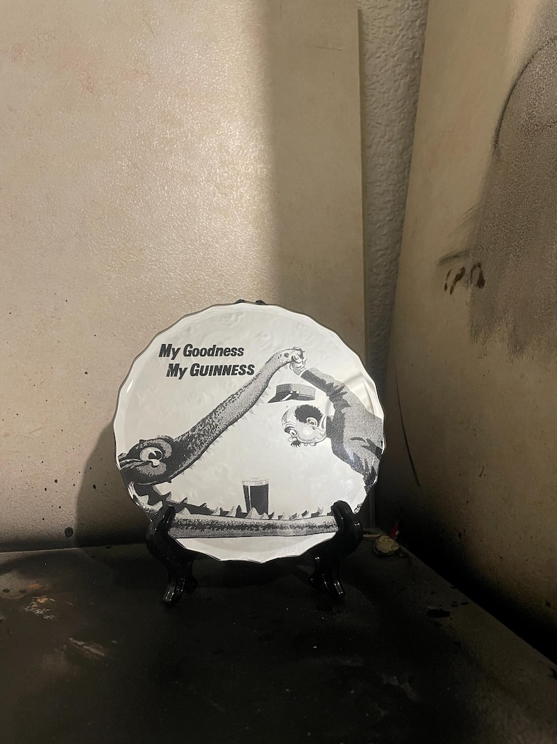 Op de afbeelding: Een zwart-wit decoratief bord met de tekst "My Goodness My Guinness". Het bord toont een cartoonillustratie van twee figuren die toosten met een pint Guinness. Het bord wordt op een zwarte standaard tentoongesteld.