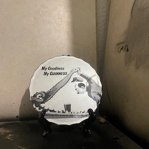 Puede incluir: Un plato decorativo redondo y blanco con una ilustración en blanco y negro. La imagen representa a dos figuras brindando sobre una pinta de cerveza, con el texto "My Goodness My Guinness". El plato está sobre un soporte negro.