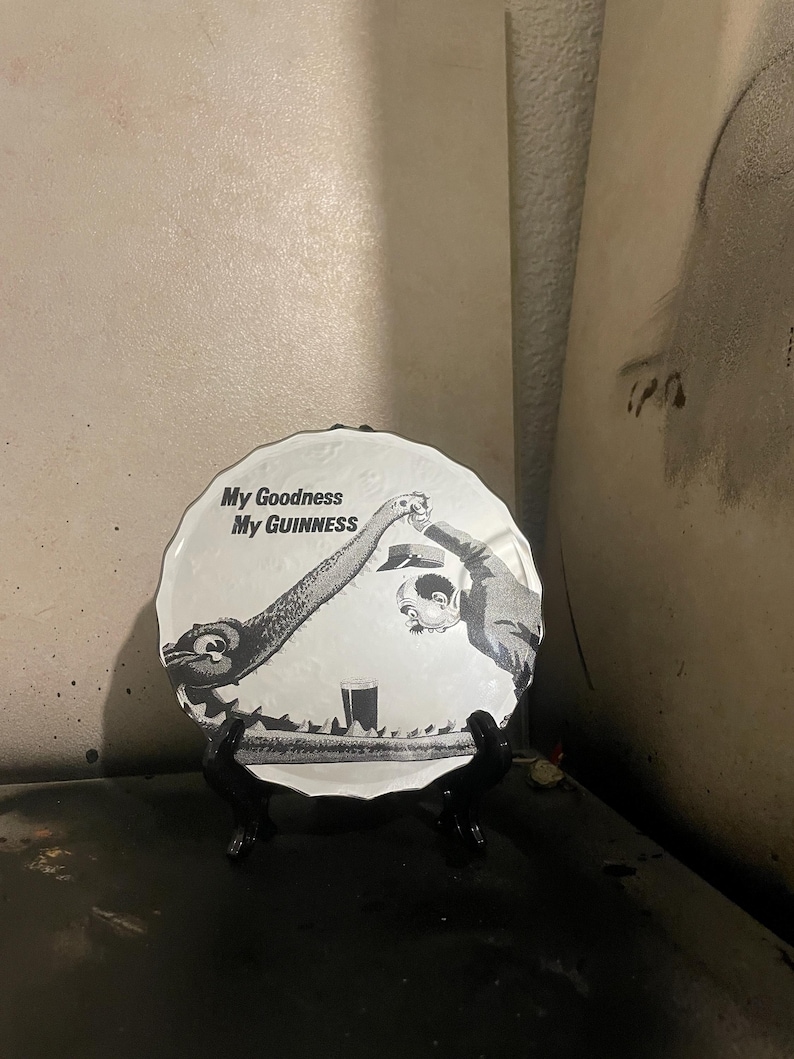 Op de afbeelding: Een ronde, witte decoratieve plaat met een zwart-wit illustratie. De afbeelding toont twee figuren die toosten op een pint bier, met de tekst "My Goodness My Guinness". De plaat staat op een zwarte standaard.