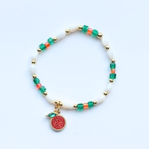 May include: A beaded bracelet featuring white teardrop-shaped beads, small green and orange cube beads, and gold-toned spherical spacers. A circular charm with a red crystal-encrusted center and a single green crystal leaf hangs from the bracelet.