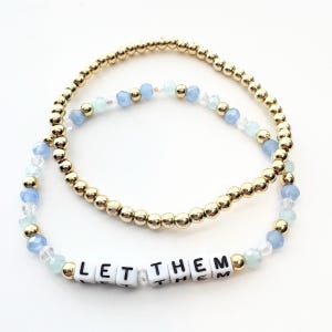 May include: Two beaded bracelets. One bracelet consists of small, round, metallic gold beads. The second bracelet features light blue and clear faceted beads interspersed with metallic gold beads and white square beads spelling LET THEM.