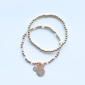 May include: Two gold-toned beaded bracelets featuring small, reflective metallic spheres and interspersed clear, iridescent beads. One bracelet includes a gold-toned charm shaped like a mouse head, encrusted with numerous small, clear crystals.