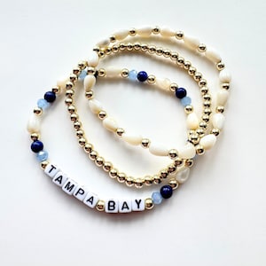 May include: Three stacked stretch bracelets featuring metallic gold beads, white teardrop-shaped beads, and dark blue and light blue round beads. One bracelet includes white square beads with black letters spelling out TAMPA BAY.
