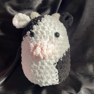 Peut inclure: Un jouet en peluche en crochet en forme de vache avec une fourrure noire et blanche. La vache a un nez rose et des yeux noirs.