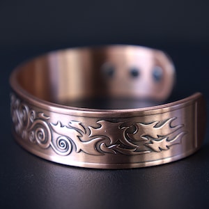 May include: A copper-colored cuff bracelet with an engraved swirling and flame-like design. The open cuff is approximately 2.5 inches wide. The bracelet has a dark background.