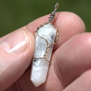 Può includere: Un ciondolo di cristallo trasparente e sfaccettato avvolto in filo d'argento e dorato. Il cristallo è bianco e grigio con una punta. Il ciondolo è tenuto in mano su uno sfondo verde sfocato.