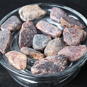 May include: A collection of small, rough, dark red and brown stones in a clear glass bowl. The stones have a natural, unpolished look.