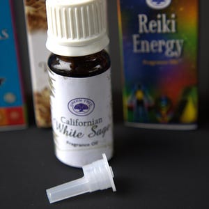 Può includere: Un piccolo flacone di vetro marrone con un tappo bianco e un'etichetta bianca che dice "Californian White Sage" e "Fragrance Oil". Il flacone è appoggiato su una superficie nera con un contagocce di plastica bianca accanto. Dietro al flacone ci sono tre scatole, una con il testo "7 Chakras", una con il testo "Reiki Energy" e una con il testo "happy yoga".