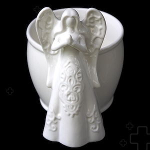 May include: A white ceramic angel figurine with folded hands, attached to a white, round planter. The angel has detailed wings and a patterned dress. The background is black.