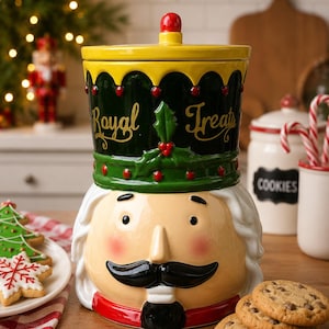 Puede incluir: Un tarro de galletas de cerámica con forma de cascanueces, tapa amarilla, sombrero verde oscuro con el texto Royal Treats en dorado, y rostro color crema con bigote negro, mejillas rojas y base de cuello rojo para guardar alimentos.