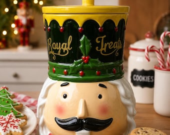 Puede incluir: Un tarro de galletas de cerámica con forma de cascanueces, tapa amarilla, sombrero verde oscuro con el texto Royal Treats en dorado, y rostro color crema con bigote negro, mejillas rojas y base de cuello rojo para guardar alimentos.