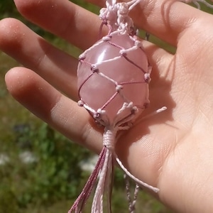 Peut inclure: Un cristal de quartz rose de couleur rose est enveloppé dans un filet de macramé avec un cordon rose et blanc. Le cristal est suspendu à un collier en cordon rose et blanc.