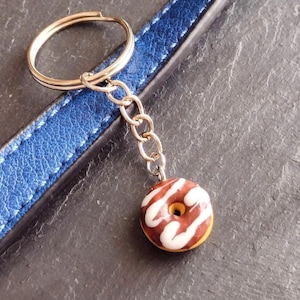 May include: A miniature donut keychain with white icing and brown stripes. The keychain is attached to a gold ring and a silver chain. A blue leather strap is in the background.